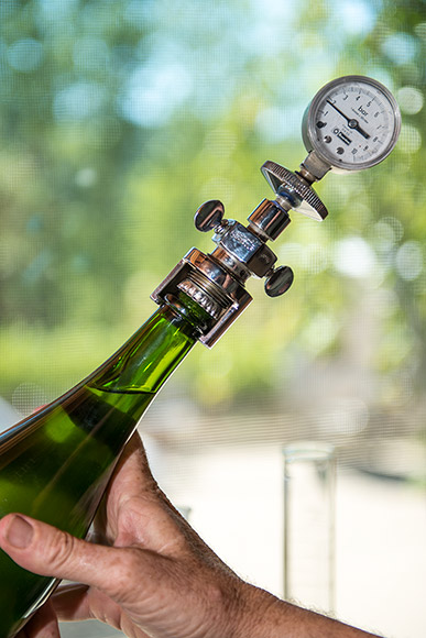 A pressure gauge pierces a crown cap on a bottle of sparkling wine.
